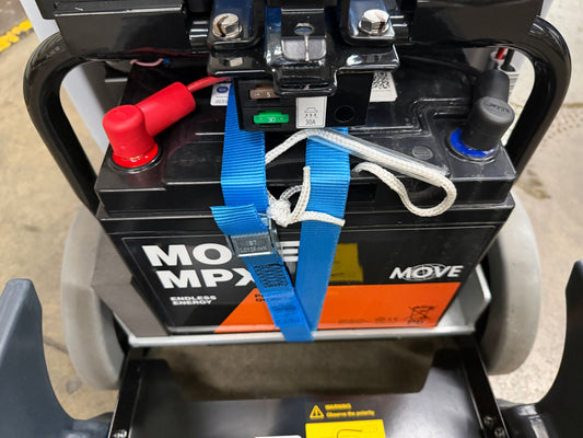 A black MOVE MPX battery secured with a blue strap, mounted on a black frame with gray wheels as part of a small walk behind floor scrubber.