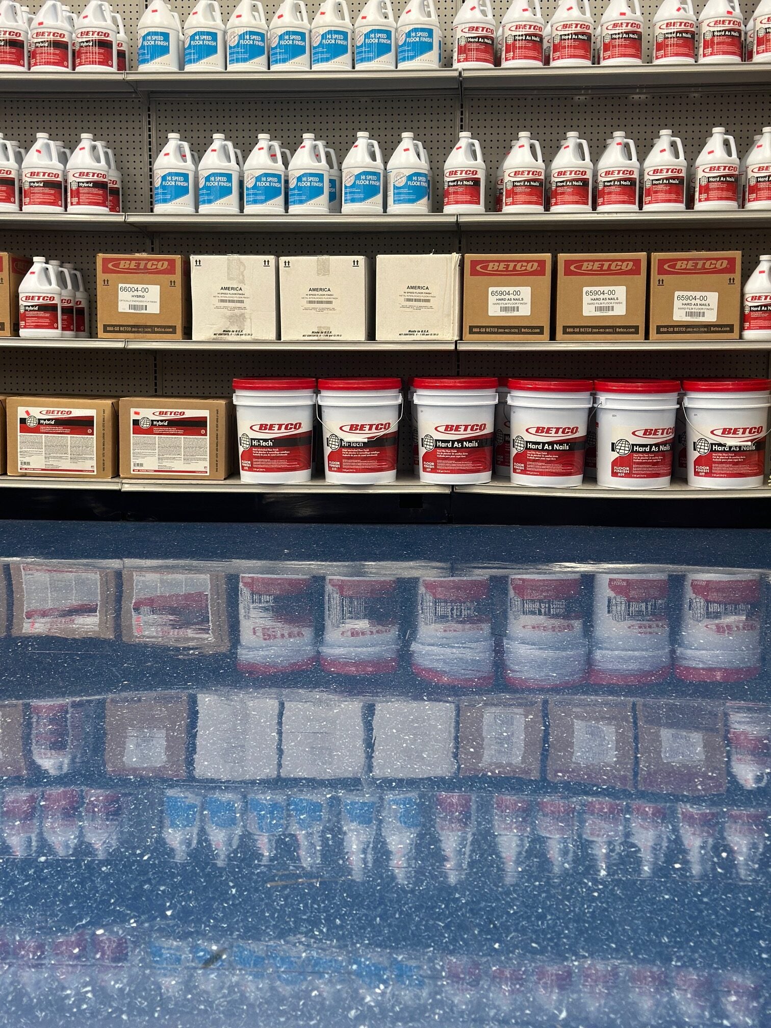 Shelves with floor finish products including bottles and boxes on a reflective floor.